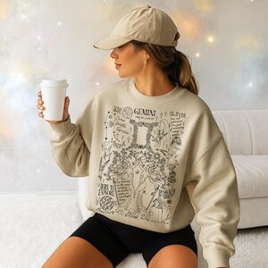 Peut inclure: Un sweat-shirt beige avec un motif zodiaque G&eacute;meaux noir. Le motif comprend les mots "Air Gemini" et "Born to Explore". Le sweat-shirt est associ&eacute; &agrave; un short noir et une casquette de baseball beige. Une tasse &agrave; caf&eacute; blanche est tenue.