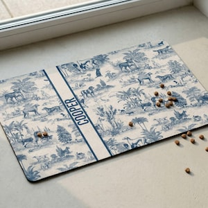 A decorative dog bowl mat with a pattern of dogs and plants, placed on a white surface near a window.