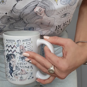 May include: White ceramic mug with a black and white illustration of an Aquarius zodiac sign. The mug has the text "Aquarius Jan 20 - Feb 18" and "Born to Envision" on it.