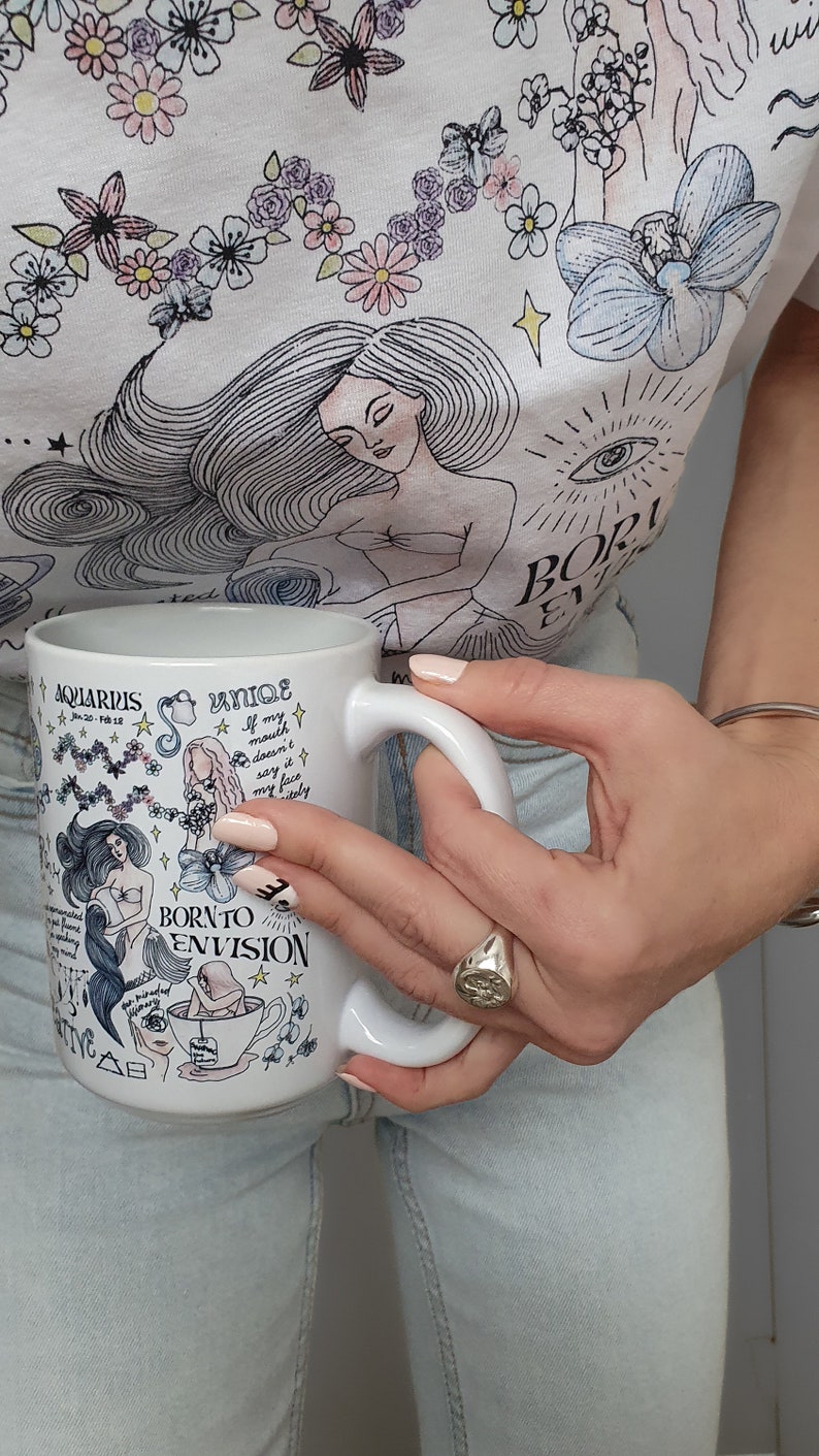 May include: White ceramic mug with a black and white illustration of an Aquarius zodiac sign. The mug has the text "Aquarius Jan 20 - Feb 18" and "Born to Envision" on it.