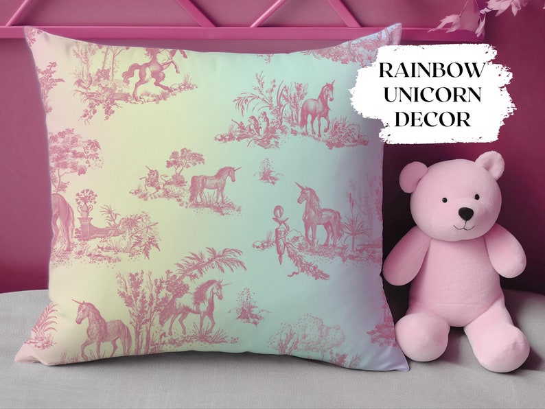 May include: A square cushion featuring a pink and white unicorn toile design. The cushion has a gradient background of yellow, green, and blue. The text "RAINBOW UNICORN DECOR" is on the cushion. A pink teddy bear is next to the cushion.