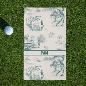May include: Golf towel with a blue and white toile design featuring golfers, golf carts, and tropical scenery. The towel has a loop for hanging and the name "PAM" printed in a horizontal band. A golf ball rests on the green grass.