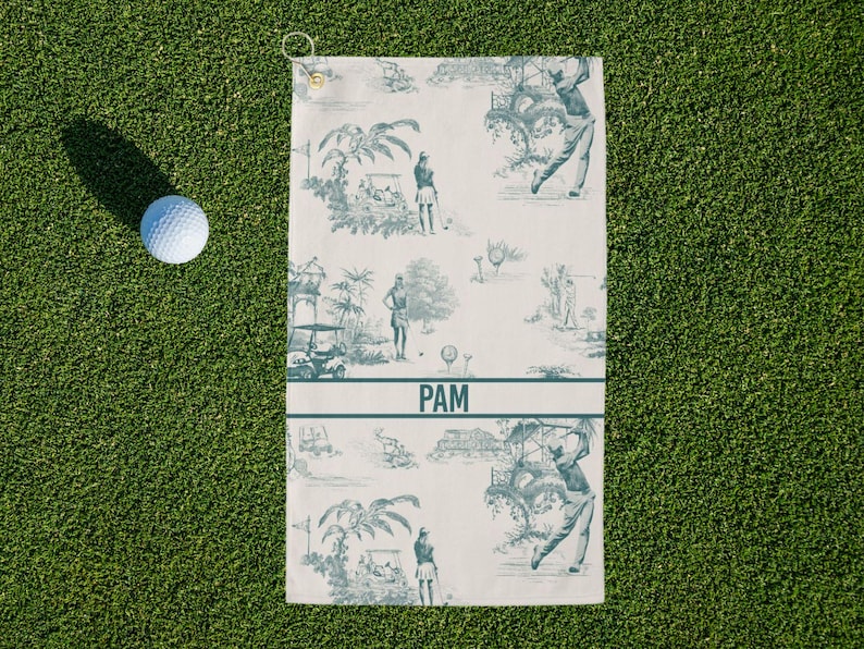 May include: Golf towel with a blue and white toile design featuring golfers, golf carts, and tropical scenery. The towel has a loop for hanging and the name "PAM" printed in a horizontal band. A golf ball rests on the green grass.
