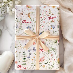 May include: A white gift box with a floral pattern and the name "Olivia" repeated on the wrapping paper. The box is tied with a beige ribbon.
