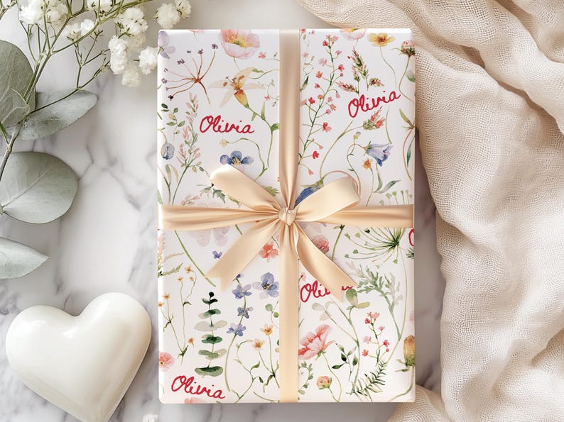 May include: A white gift box with a floral pattern and the name "Olivia" repeated on the wrapping paper. The box is tied with a beige ribbon.