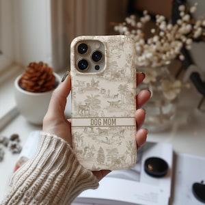 Op de afbeelding: Een witte iPhone-hoes met een beige en bruin toile-patroon met honden en bomen. De hoes heeft een gouden band met de tekst "DOG MOM" erop gedrukt.