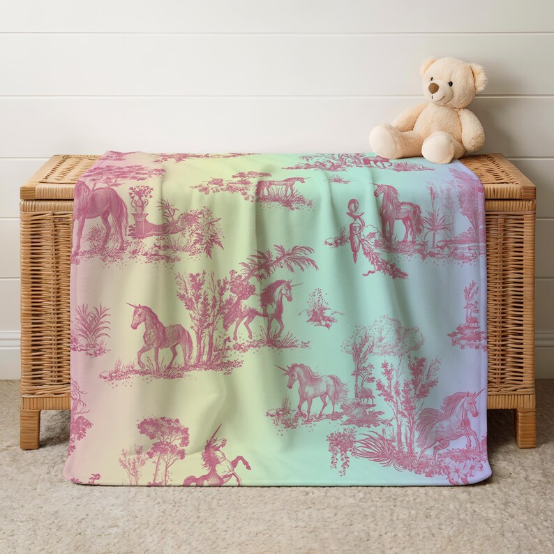 May include: A soft blanket with a pink unicorn toile pattern on a pastel rainbow gradient background. The blanket is draped over a wicker chest, with a teddy bear on top. The blanket features unicorns, trees, and other pastoral elements.