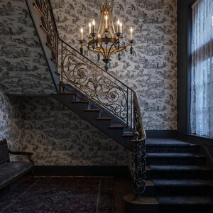 May include: An interior shot of a staircase with a decorative wrought iron railing and a chandelier. The walls are covered in a black and white toile wallpaper. A vintage bench sits at the base of the stairs, and a window with sheer curtains is visible.