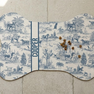 A bone-shaped dog food mat with a pattern of various animals and plants, placed on a tiled floor.