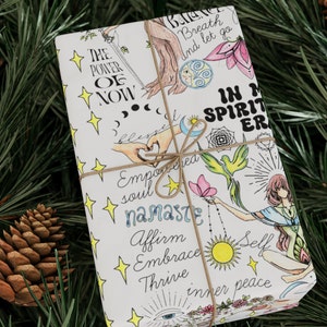 May include: White gift wrap with a colorful design featuring illustrations of hands, a butterfly, a bird, a sun, a moon, stars, and flowers. The text on the wrapping paper reads "The Power of Now", "Breath and let go", "In My Spirit", "Empowered Soul", "Namaste", "Affirm", "Embrace", "Thrive", "Self", and "Inner Peace".