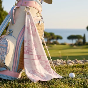 May include: A golf bag with a matching towel, featuring a pink and white design. The towel is personalized with the name "OLIVIA". The bag has blue and pink accents, with a golf ball on the grass in the foreground.