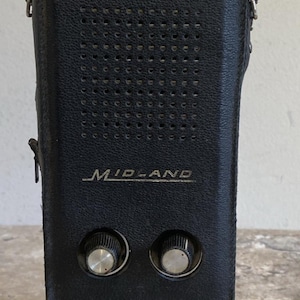 以下が含まれることがあります： シルバーのアンテナが付いた黒いヴィンテージMidlandトランシーバー。このデバイスには、2つのシルバーノブ、スピーカーグリル、Midlandのロゴが付いています。前面には小さな白いステッカーが付いています。トランシーバーは黒い革ケースに入っています。