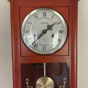 May include: A rectangular wooden wall clock with a warm brown finish, featuring a round white face with black Roman numerals and the text KASSEL and 15 DAY. The lower section has a glass panel displaying a gold-coloured pendulum with floral designs.