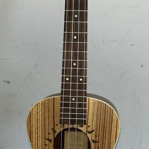 May include: A wooden ukulele with a sun design around the sound hole. The instrument has a striped wood grain pattern and a dark brown fretboard. The headstock has tuning pegs.