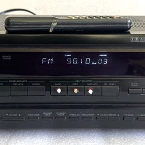 Pode incluir: Receptor de áudio/vídeo Sherwood preto com um controle remoto em cima. O receptor tem um display digital mostrando "FM 98.10.03". Possui um botão de energia, controle de volume e várias tomadas de entrada/saída. O receptor é rotulado "HD-6300".