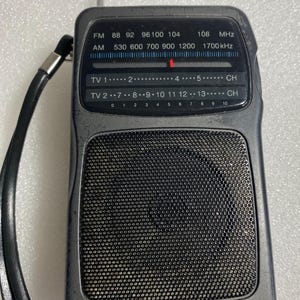 May include: A black portable Radio Shack AM/FM radio with a metal antenna, a black wrist strap, and a front speaker grille. The display panel shows frequency scales for FM, AM, and TV channels with a red indicator needle and white text markings.