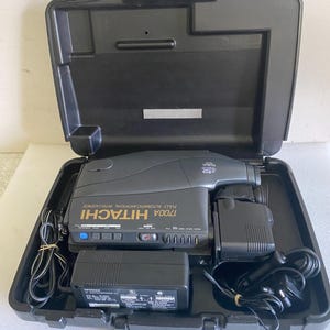 Puede incluir: Una videocámara Hitachi 1700A negra con el texto Fully Automatic Artificial Intelligence, guardada en un maletín de transporte de plástico negro. El conjunto incluye un adaptador de corriente negro y dos cables de alimentación negros, organizados en el interior.