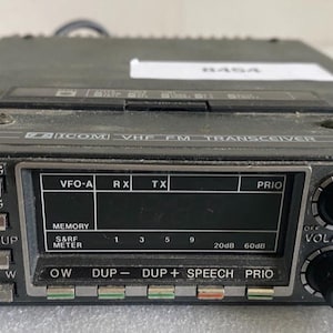 Puede incluir: Un transceptor VHF FM Icom IC-27A negro con pantalla digital, múltiples botones pulsadores etiquetados como M/VFO, A/B, S/S, TS, 1M UP, M W, HI/LO, TONE, OW, DUP-, DUP+, SPEECH y PRIO, junto con dos perillas de control negras y conector.