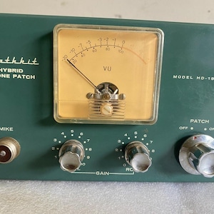 Pode incluir: Um dispositivo Heathkit Hybrid Phone Patch modelo HD-19 verde-escuro com um medidor VU analógico central de fundo bege, dois botões de controle de ganho, um interruptor de patch on-off e uma entrada de microfone marcada como MIKE.