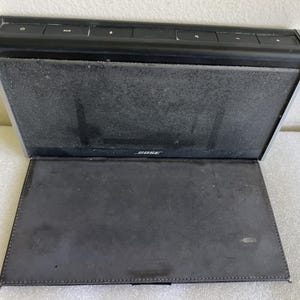 May include: A black Bose portable speaker with a dark grey mesh speaker cover. The speaker has a rectangular shape with a silver trim and a built-in cover. The Bose logo is visible on the speaker.