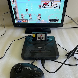 Może przedstawiać: Czarna konsola do gier Sega Genesis z podłączonym kontrolerem i kartridżem do gry w hokeja. Ekran telewizora wyświetla grę w hokeja z zawodnikami w czerwonych i białych strojach. Gra jest oznaczona jako "Hockey".
