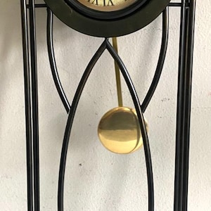 May include: A tall wall clock with a black metal frame featuring an arched top and curved vertical supports. The clock face has a gold-toned background with black Roman numerals and the text "Cateau Clocks". A gold-colored pendulum hangs below.