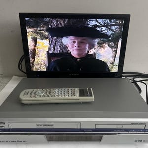 Pode incluir: Um videocassete prateado e preto com controle remoto está posicionado abaixo de uma TV de tela plana preta. A TV exibe uma cena de um filme. O videocassete tem as palavras "Hi-Fi Stereo" impressas na frente.