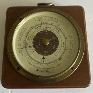 May include: A square wooden barometer with a circular brass-rimmed face. The cream-colored dial features black text including 'Airguide', 'Stormy', 'Rain', 'Fair', and 'Dry', with a central brown altitude scale and a small metal hanging loop at the top.