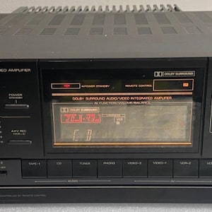 May include: A black Fisher Studio Standard AV surround stereo amplifier featuring a central digital display, sliding bass and treble controls on the left, and volume and balance controls on the right. The front panel includes various input buttons.