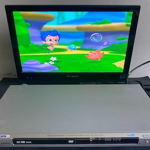 May include: A silver Sony DVD player with an HDMI port sits in front of a black Dynex television screen. The television displays a colourful animated scene featuring a blue-haired character, a small orange fish, and a blue creature in a green landscape.