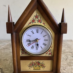 May include: A wooden mantel clock with a decorative floral design. The clock has a white face with black Roman numerals and gold trim. The clock is housed in a brown wooden frame with two pointed finials.