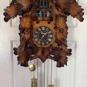 May include: A wooden cuckoo clock featuring carved grape leaves and birds, a black circular dial with gold Roman numerals, and the text The Timco Quartz. Two small figures stand on a balcony above the dial, with two brass weights hanging below.