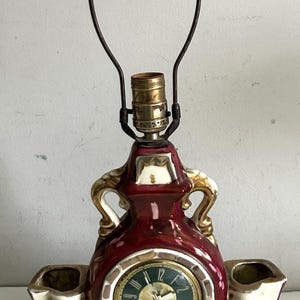 May include: A vintage burgundy ceramic lamp with a clock face and two small vases. The lamp has gold accents and floral details. The clock face is green with gold numerals. The lamp is a decorative home decor item.
