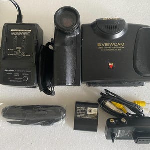 Puede incluir: Una videocámara de cristal líquido Sharp Viewcam modelo VL-E47 negra con un cargador de batería, una batería negra, una correa de hombro negra en embalaje de plástico y un cable AV negro con conectores amarillos y blancos para salida de audio y vídeo.