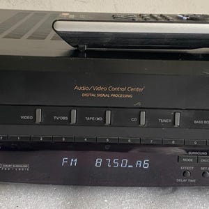 May include: A black Sony Audio/Video Control Centre receiver with a digital display showing FM 87.50 A6. The front panel features a large master volume knob, input selection buttons, a headphone jack, and a remote control resting on top.