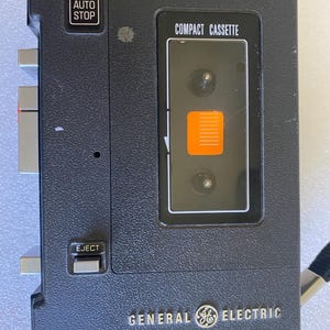 Peut inclure: Un enregistreur de cassettes portable General Electric gris foncé doté d'un compartiment à chargement par le haut marqué COMPACT CASSETTE avec un indicateur orange. L'appareil comprend des boutons de commande argentés, un interrupteur EJECT et les mentions ALC et AUTO STOP.