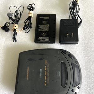 Puede incluir: Un reproductor de CD portátil Memorex DASP, una cinta de cassette, un adaptador de corriente y auriculares. El reproductor de CD es de color gris oscuro con una pantalla digital y botones de control. La cinta y el adaptador son negros.