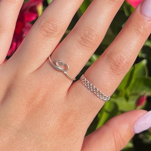 May include: A hand with white ombre nails and two silver rings. The hand is in front of a background of pink flowers and green leaves.