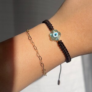 May include: A brown string bracelet with a blue evil eye charm in the shape of a flower. The bracelet is tied with a knot and has a tassel.