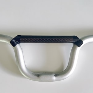 Könnte beinhalten: Silberner Fahrradlenker mit einem schwarzen Mittelteil aus Kohlefaser. Der Lenker hat ein gebogenes Design, wahrscheinlich für Mountainbikes. Der Lenker wird vor einem weißen Hintergrund gezeigt. Die Breite des Lenkers beträgt ca. 60 cm.