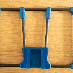 May include: A blue and black rectangular frame with a unique design. The frame has blue corner pieces and a central blue component, connected by black rods. The object is set against a light wood grain background.