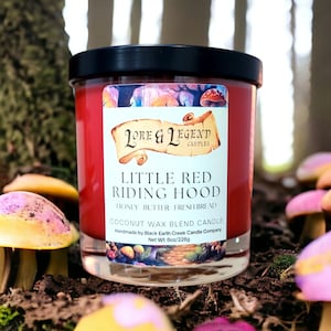 May include: A red candle in a glass jar with a black lid. The label reads "LORE & LEGEND CANDLES LITTLE RED RIDING HOOD HONEY BUTTER FRESH BREAD COCONUT WAX BLEND CANDLE Handmade by Black Earth Creek Candle Company Net Wt: 8oz/226g".