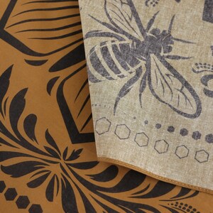 May include: A brown fabric with a black floral pattern and a honeycomb design. A beige fabric with a black bee design is layered on top.