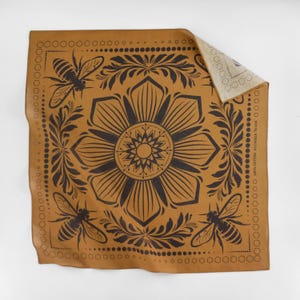 May include: A brown leather bandana with a black floral design and bee motifs. The bandana has a repeating pattern of bees, flowers, and leaves. The bandana is folded in half and the back side is visible.