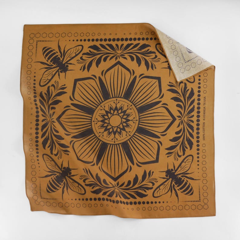 May include: A brown leather bandana with a black floral design and bee motifs. The bandana has a repeating pattern of bees, flowers, and leaves. The bandana is folded in half and the back side is visible.