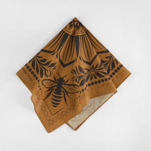 May include: A brown bandana with a black bee design and a floral pattern. The bandana has a white border and the text "HANDMADE TX USA" is printed on the border.