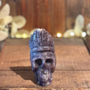 May include: A carved amethyst skull with a crown-like design on top. The skull is a deep purple color with lighter streaks and sits on a wooden surface. The background is blurred, with hints of light and foliage.
