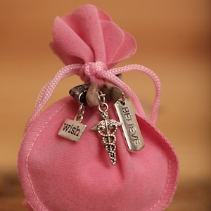 May include: A pink velvet pouch with a drawstring closure and silver charms. The charms have the words "wish" and "believe" on them. The pouch is sitting on a wooden surface.