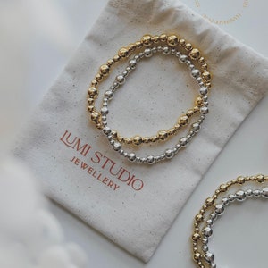 Könnte beinhalten: Zwei Perlenarmbänder, eines golden und eines silbern, liegen auf einem weißen Stoffbeutel. Der Beutel trägt den Aufdruck "Lumi Studio Jewellery".