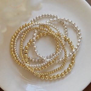 May include: Several beaded bracelets in gold and silver are displayed on a white plate. The bracelets are made of small, spherical beads, offering a refined and graceful appearance. The image is well-lit, emphasizing the metallic lustre of the jewellery.
